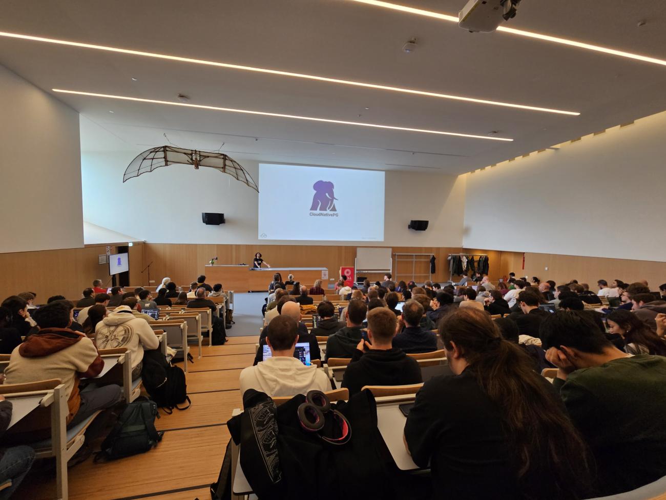 Boriss Mejias presenting his PGConf BE session to a packed room, the CloudNativePG logo on a slide